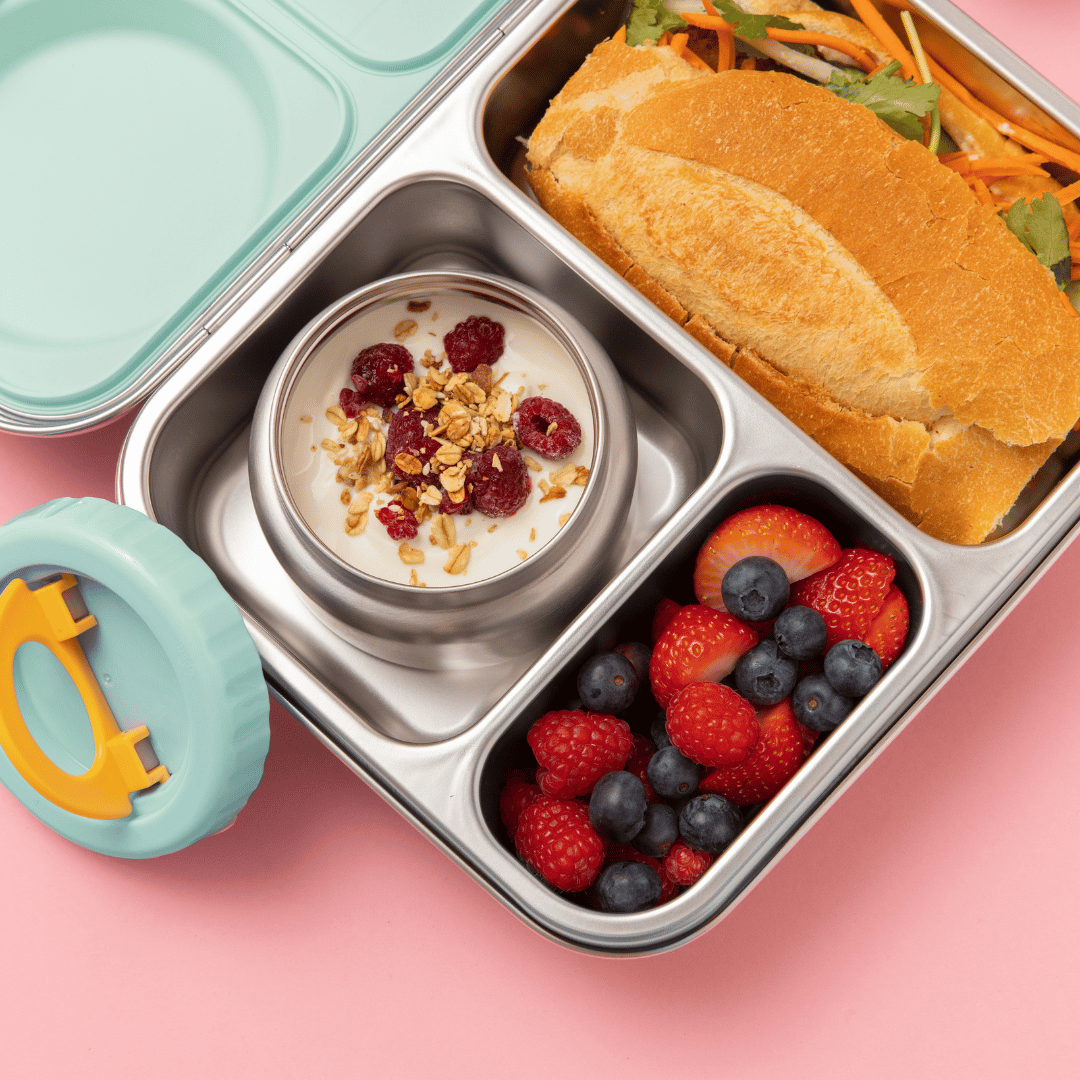 Hot n Cold stainless steel hero lunch box box in mint includes an insulated food jar that sits inside with 2 other compartments containing a sandwich, fruit, and other snacks on a pink background