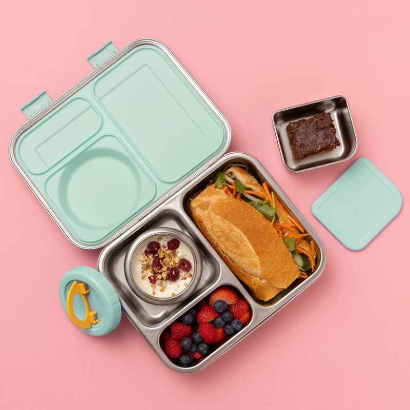 Hot n Cold stainless steel hero lunch box box in mint includes an insulated food jar that sits inside with 2 other compartments containing a sandwich, fruit, and other snacks on a pink background
