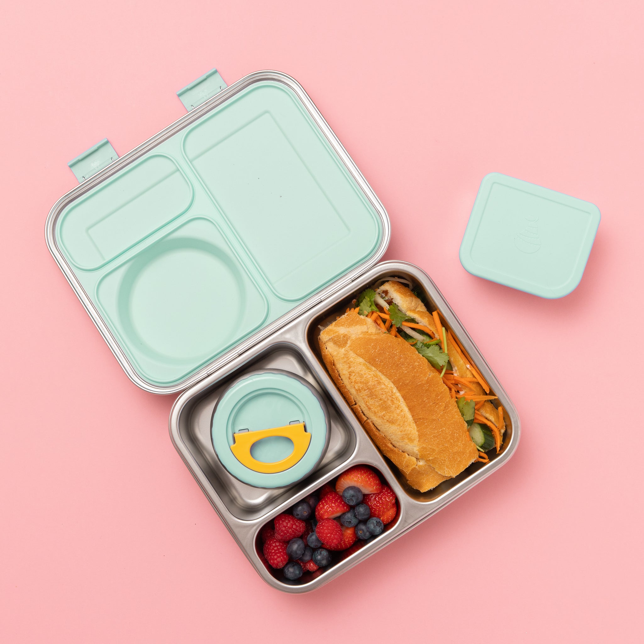 Hot n Cold stainless steel hero lunch box box in mint includes an insulated food jar that sits inside with 2 other compartments containing a sandwich, fruit, and other snacks on a pink background