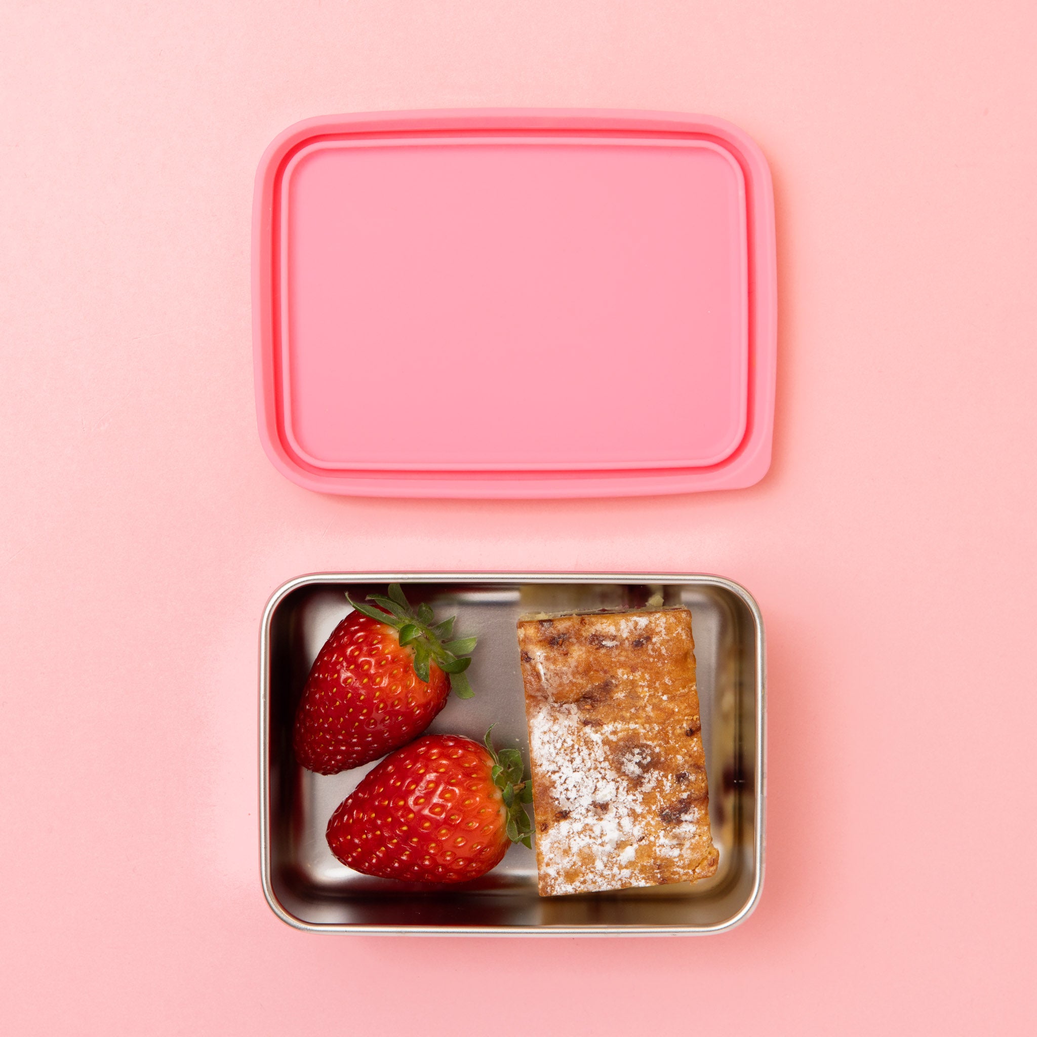 stainless steel snack container with pink silicone lid on a pink background with a slice and strawberries