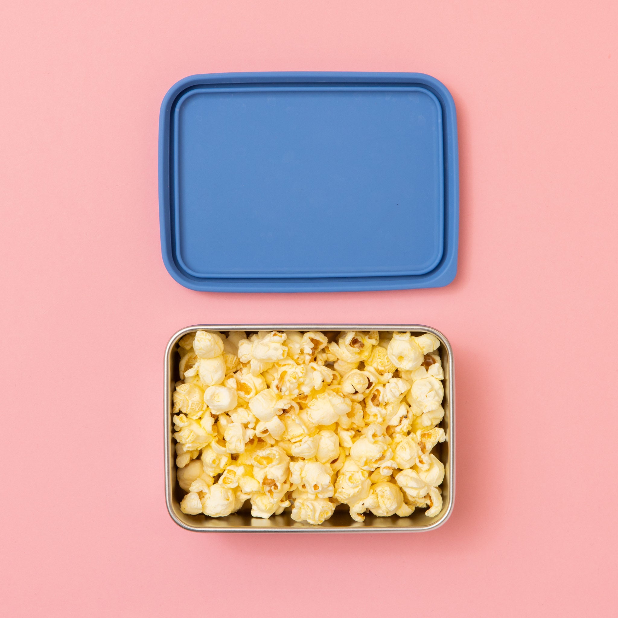 rectangular stainless steel snack container with popcorn and blue lid on a pink background