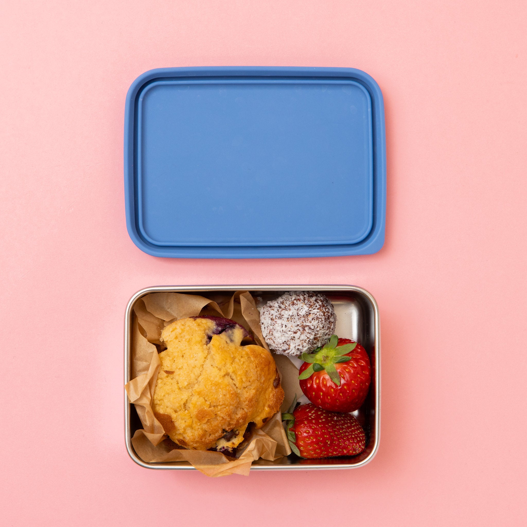 rectangular stainless steel snack container with a muffin bliss ball and strawberries and blue lid on a pink background
