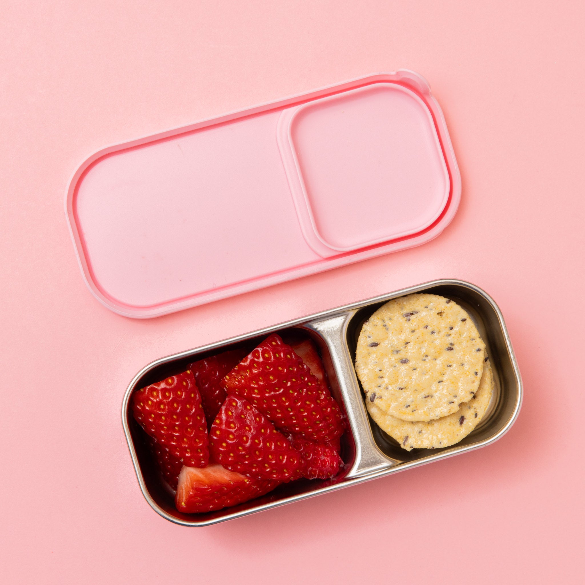 two compartment stainless steel snack container with light pink silicone lid shown with strawberries and rice cracker 