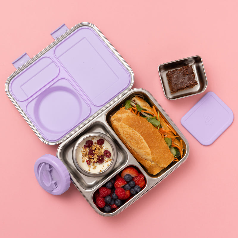 A stainless steel bento lunch box with a lavender purple lid, shown open on a solid pink background. The lunch box is filled with a sandwich, yogurt with berries, and a small side dish, with its separate lavender lid and small square container placed nearby.